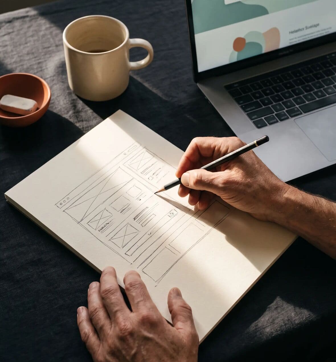 Mãos a desenhar wireframes de um site ao lado de um portátil com o design final